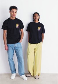 Sorte bomulds t-shirts med farverige grafiske tryk på venstre bryst, sammen med lyseblå jeans og gule vidde bukser.