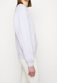 Sweatshirt lavande clair avec poignets et ourlet côtelés, présentant des détails de couture sur les coutures latérales. Le modèle est porté avec un pantalon de couleur claire.