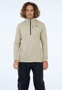 Beżowy sweter z półzamkiem i długimi rękawami, z teksturowanym wykończeniem oraz małym logo na piersi. Noszony z czarnymi spodniami.