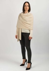 Beige uldeskaut, der hænger over en hvid top, parret med sorte skinny jeans og sorte højhælede sko. Glat tekstur, elegant drapering.