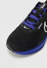 Scarpa sportiva nera con tomaia in mesh, lacci viola, dettagli bianchi e suola blu. Presenta una punta arrotondata e un materiale testurizzato.