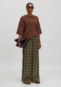 Übergroßer brauner Pullover, karierte grüne Maxiröcke mit orangefarbenen und blauen Linien, schwarze Clutch-Tasche, braune Slipper, Sonnenbrille.