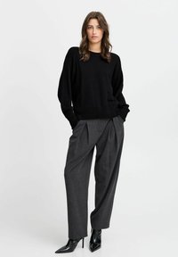 Schwarzer gerippter Strickpullover mit oversized Passform, kombiniert mit hochtaillierten grauen, gefalteten Hosen und spitzen schwarzen Stilettos.