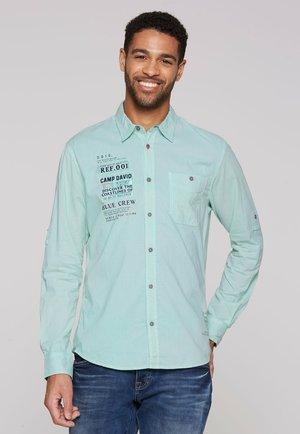 Camisa de manga larga verde claro con botones en la parte delantera, un bolsillo en el pecho y gráficos impresos en negro. Presenta botones en contraste y una textura de tela diferente.