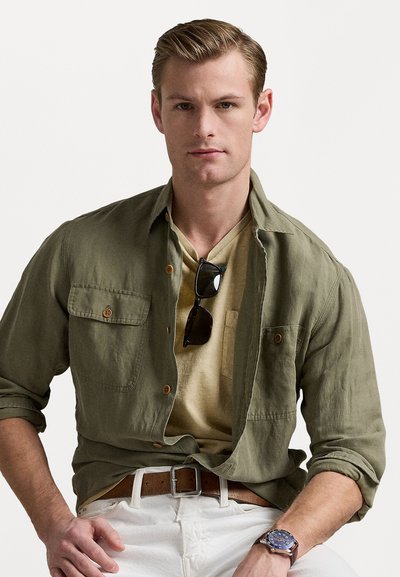 Αρκετά ελαφρύ ελαιόχρωμο πουκάμισο με κουμπιά και δύο τσέπες στο στήθος, φορεμένο πάνω από ένα μπεζ T-shirt, σε συνδυασμό με λευκά παντελόνια και καφέ ζώνη.