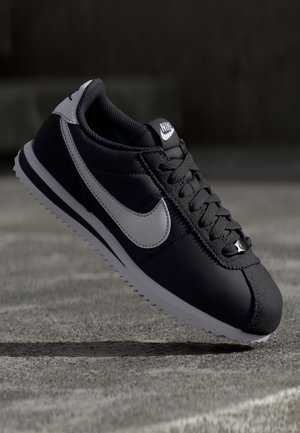 Czarno-biały but sportowy Nike Cortez z sznurowadłami, białym logo Swoosh i ryflowaną podeszwą na betonowej powierzchni z ciemnym, rozmytym tłem.