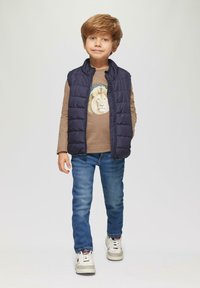 Marine gewatteerde vest over een bruine lange mouw shirt met een leeuwengrafiek, gecombineerd met blauwe jeans en witte sneakers. Ongelijke texturen en kleuren aanwezig.