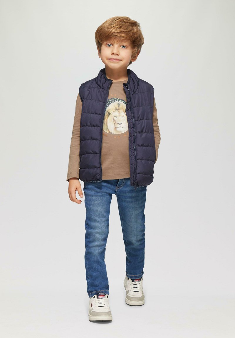Marine gewatteerde vest over een bruine lange mouw shirt met een leeuwengrafiek, gecombineerd met blauwe jeans en witte sneakers. Ongelijke texturen en kleuren aanwezig.