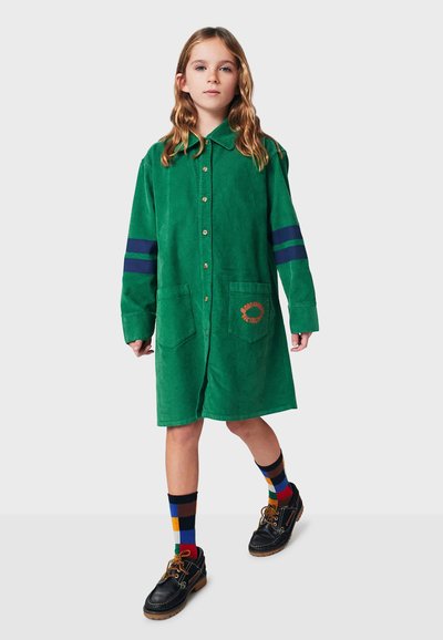 Grünes Cordhemdkleid mit Knopfleiste, zwei großen Taschen und dunkelblauen Streifen an den Ärmeln. Trägt bunte Socken und schwarze Schuhe.