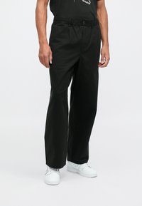 Sorte, løstsittende bukser med elastisk midje og knappelukking, laget av glatt stoff, sammen med hvite joggesko.