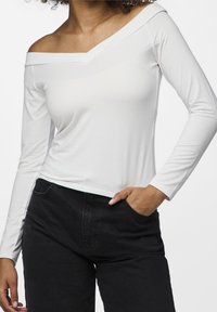 Vit långärmad topp med off-shoulder-design och slät textur, kombinerad med mörka högmidjade jeans. Har en figurnära siluett.