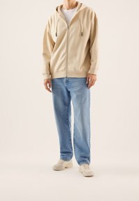 Man in een beige ritsvest, lichtblauwe jeans, wit t-shirt en beige sneakers, staand tegen een effen lichte achtergrond.