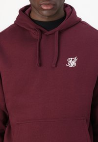 Sweat à capuche bordeaux en mélange de coton doux, avec une poche kangourou à l'avant et un logo brodé en blanc sur la poitrine.