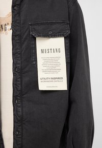 Black denim shirt with a button-down design, featuring a chest pocket and a hanging tag with product details. Soft, durable fabric.
