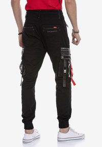 Schwarze Cargo-Hosen mit mehreren Taschen, elastischen Bündchen und dekorativen Hardware-Akzenten. Das Material wirkt langlebig mit einem matten Finish.