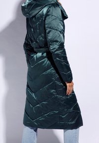 Abrigo de plumas largo en un verde oscuro con un patrón de chevron, capucha y cintura ceñida con cinturón. Tela lisa y brillante con un diseño hasta la rodilla.