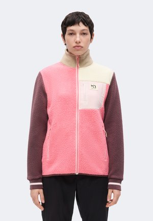 Personne portant une veste polaire color-block rose, crème et violette avec une fermeture éclair devant et une poche poitrine, debout devant un fond uni.