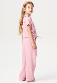 Roze tweedelig outfit gemaakt van textiel met structuur. Korte mouwen top met kraag, strik rond de taille en wijde broekspijpen. Schoenen met een veelkleurig patroon.