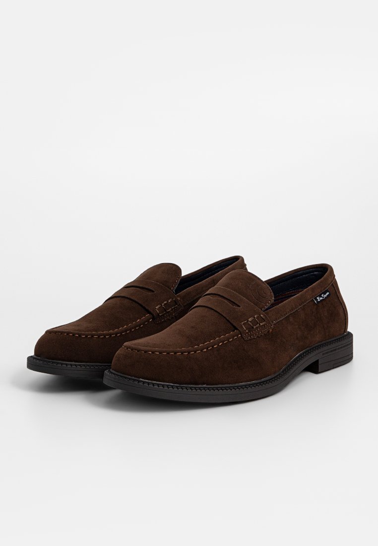 Mocasines de ante marrón con costura visible, suela de goma plana y una pequeña etiqueta de logo en el costado. Diseño clásico con una silueta elegante.