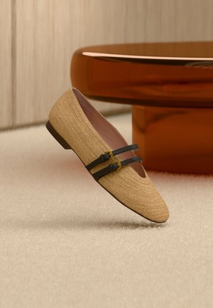 Μπεζ υφαντό επίπεδο παπούτσι με δύο μαύρες λουρίδες και χρυσές πόρπες, τοποθετημένο σε φωτεινό χαλί με φόντο καφέ στρογγυλό τραπέζι.