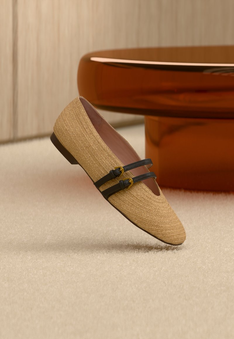 Μπεζ υφαντό επίπεδο παπούτσι με δύο μαύρες λουρίδες και χρυσές πόρπες, τοποθετημένο σε φωτεινό χαλί με φόντο καφέ στρογγυλό τραπέζι.