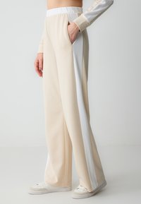 Beige sweatpants met witte zijkanten, wijdvallend ontwerp, elastische tailleband en zijzakken. Gecombineerd met witte sneakers.