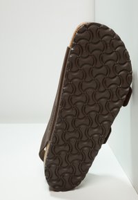 Calzado marrón con una suela de goma texturizada que presenta patrones circulares. El material superior parece suave con un diseño simple.