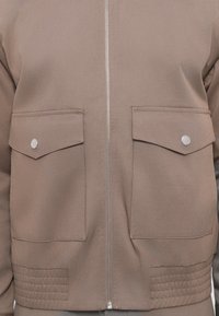Zip-up beige jacket with two front flap pockets secured by metal snaps and stitched detailing at the bottom hem.