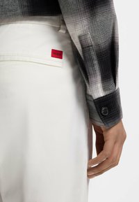 Μακρυμάνικο πουκάμισο σε ασπρόμαυρο καρό με μανσέτα με κουμπιά, συνδυασμένο με λευκό παντελόνι που φέρει κόκκινη ετικέτα "HUGO" στην πίσω τσέπη.