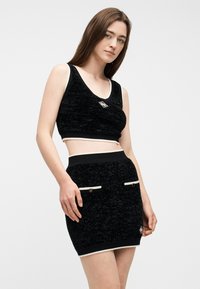 Νεαρή γυναίκα που φοράει μαύρο υφασμάτινο αμάνικο crop top και αντίστοιχη ψηλόμεση μίνι φούστα με λευκή επένδυση και μπροστινές τσέπες.