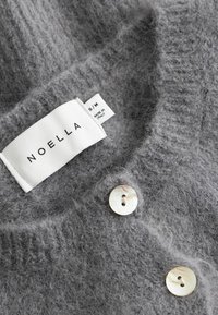 Weicher grauer Strickstoff mit zwei perlmuttfarbenen Knöpfen und einem weißen Kleidungsetikett mit der Aufschrift "NOELLA" sowie der Größe "S/M Hergestellt in Italien."