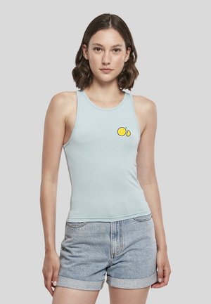 Jeune femme portant un débardeur bleu clair avec un motif de tranche de citron et un short en jean retroussé, debout devant un fond uni.