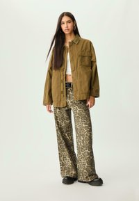 Žena s dugom tamnom kosom koja nosi preveliku maslinastu jaknu, bijeli kratki top, široke hlače s leopard printom, crni remen i crne cipele stoji.