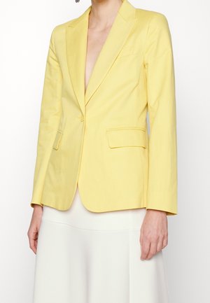 Personne portant un blazer ajusté jaune vif avec des poches à rabat sur une jupe blanche, sur un fond blanc uni.