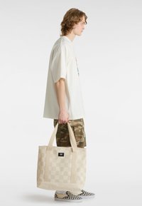 Borsa a spalla a scacchi beige con doppi manici, realizzata in tessuto resistente. Indossata da una persona con una camicia bianca larga e pantaloni corti mimetici.
