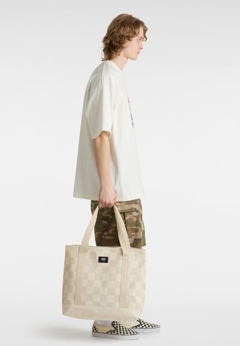 Borsa a spalla a scacchi beige con doppi manici, realizzata in tessuto resistente. Indossata da una persona con una camicia bianca larga e pantaloni corti mimetici.