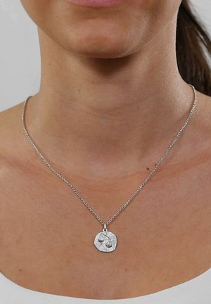 Silver chain necklace with a round pendant featuring a scales of justice symbol, worn on a person's neck above a white top.