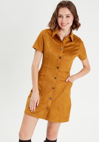 Robe à manches courtes en faux daim moutarde. Elle possède un col, des poches à l'avant et des détails de couture verticaux. Texture lisse sur l'ensemble.