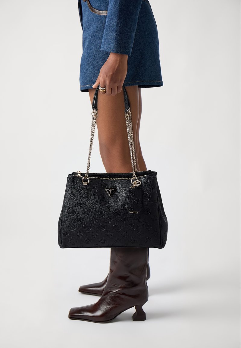 Personne en jupe bleue tenant un sac à main noir texturé avec des anses chaîne, portant des bottes marron mi-mollet à talons sur fond blanc.