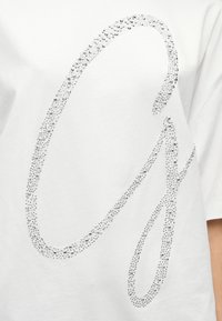 Λευκό μπλουζάκι με μεγάλο, λαμπερό ασημένιο σχέδιο από στρας σε καλλιγραφικό στυλ, με λεία υφή υφάσματος και κοντά μανίκια.