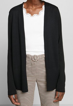 Personne portant un cardigan noir ouvert, un haut blanc avec un col en dentelle et un pantalon à carreaux taille haute avec une boucle de ceinture circulaire.
