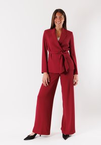 Blazer tailored borgogna con vita legata, abbinato a pantaloni larghi coordinati, realizzati in tessuto morbido, con revers affilati e dettagli minimali.