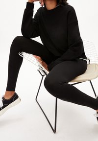 Czarny sweatshirt i legginsy, siedząca na drucianym krześle. Białe sneakersy z czarnymi akcentami. Gładki materiał, dopasowany krój, casualowy wygląd.