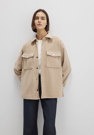 Beige wool button-up shirt jacket with two chest pockets, a pointed collar, and a relaxed fit, paired with dark blue jeans.