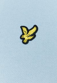 El tejido de punto azul claro presenta un logo de pájaro amarillo bordado contorneado en negro. El diseño está centrado en la suave textura del material.
