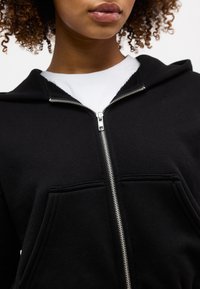 Κοντινό πλάνο ενός ατόμου που φοράει μαύρη ζακέτα με φερμουάρ πάνω από λευκό πουκάμισο, εμφανίζοντας το φερμουάρ και τις μπροστινές τσέπες.