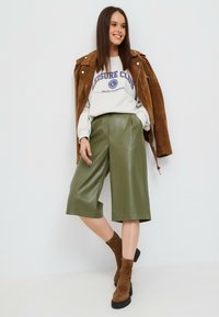 Oliwkowe kuloty z faux skóry zestawione z jasnoszarym sweatshirtem z granatowym napisem, uzupełnione brązową zamszową kurtką i botkami za kostkę.