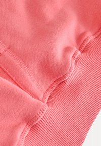 Koralni pulover z mehko teksturo, rebrastimi manšetami in gladkimi šivi. Tkanina je videti puhasta in topla z enobarvnim dizajnom.