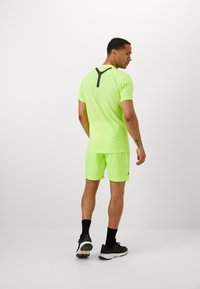 Camiseta y pantalones cortos deportivos de color verde neón, con una textura de malla y detalles negros en la parte posterior. Combinados con zapatillas negras y calcetines hasta el tobillo.