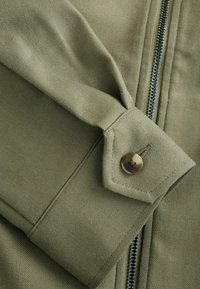 Olivgrüne Jacke mit Fischgrätmuster, Reißverschluss und Knopfdetails an den Ärmelbündchen. Der Stoff hat eine glatte Oberfläche.